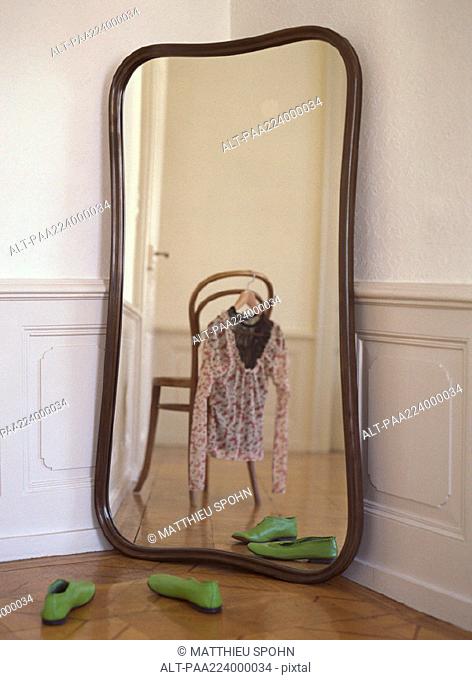 Mirror reflecting chair with blouse hanging from it and pair of shoes on floor