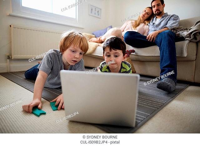 Brothers playing laptop game on room rug, parents on sofa