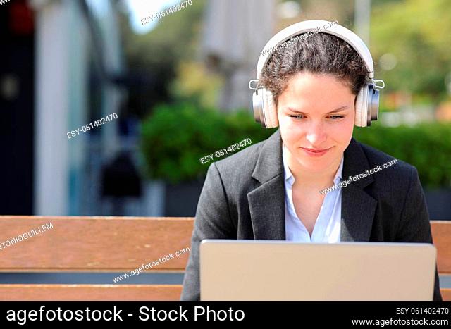 Businesswoman wearing wireless headphones using laptop sitting on bench