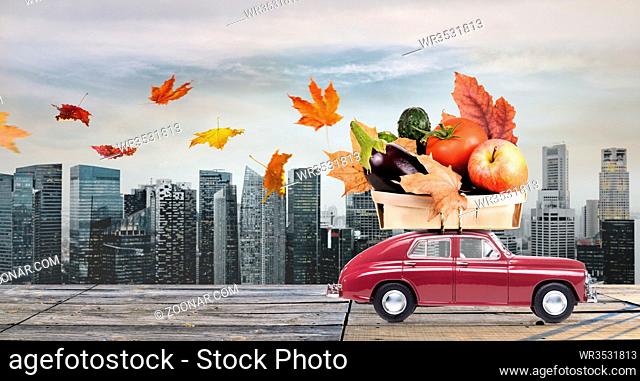 Food delivery. Autumn red toy car with fallen leaves delivering fruits and vegetables against business district buildings