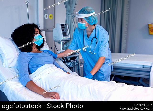 Caucasian female doctor in hospital in face mask consoling female patient in bed wearing face mask