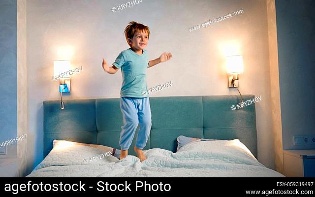 Happy laughing and smiling toddler boy in pajamas skipping and jumping up high on parent's bed at night before going to sleep