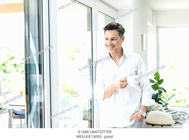 Mature man leaning on window, drinking coffee at home
