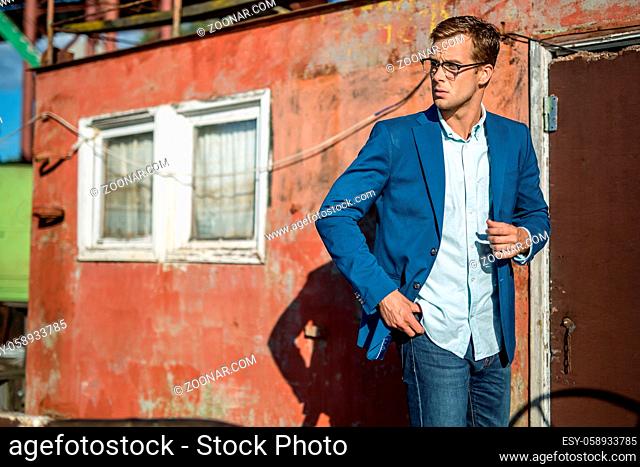 Stylish man in glasses stands outdoors on the decrepit building background. He wears blue jeans, a light shirt and a blue jacket