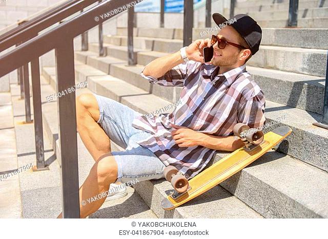 Cheerful male skater is talking on mobile phone and laughing. He is sitting on stairs outdoors and relaxing