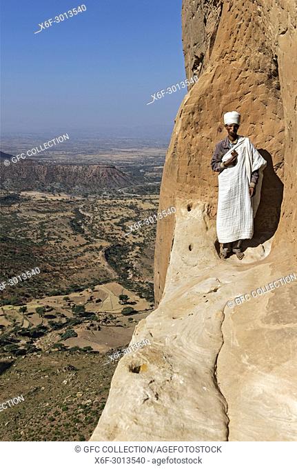 Priest standing on an exposed rock ledge at the entry to the rock-hewn church Abuna Yemata high above the ground, Gheralta region, Tigray, Ethiopia