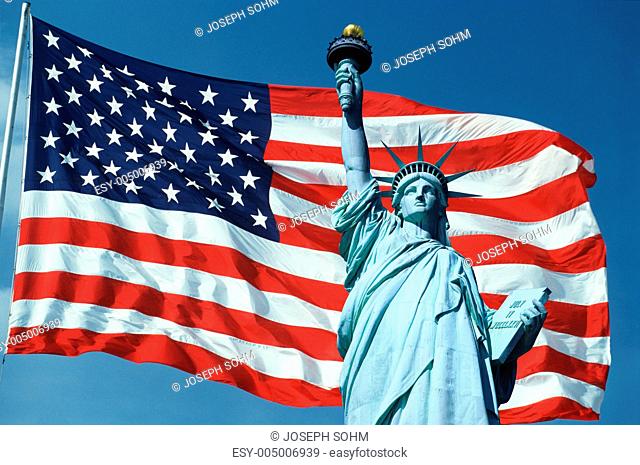 This is an American flag on a flagpole waving in the wind set against a blue sky. The top half of the Statue of Liberty is digitally composited onto the right...
