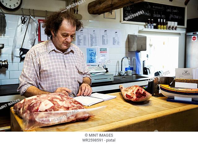 Butcher standing at the counter of his shop, looking at a notebook