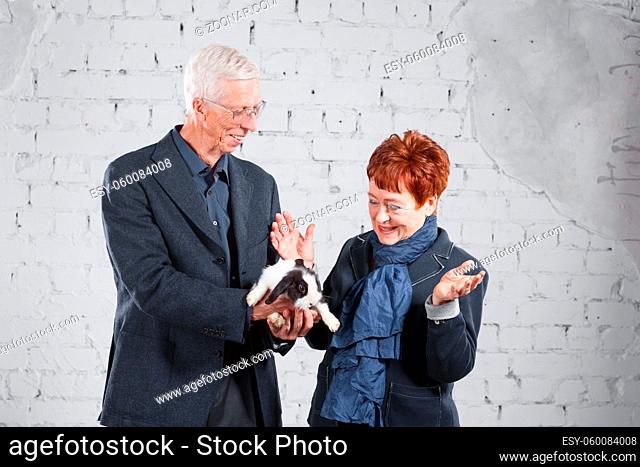 Happy smiling old couple standing together with pet rabbit on white brick background