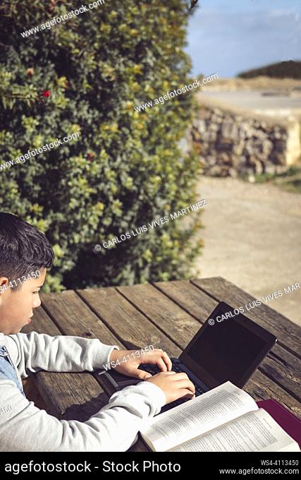 young latin student, outdoors, using laptop, sitting at a park table working with books and computer