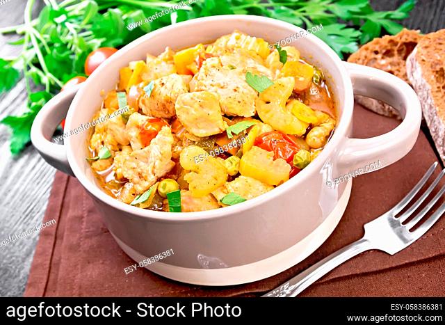 Stew of chicken breast, tomatoes, stalked celery, carrots, green peas and onions in a tureen on brown napkin on dark wooden board background