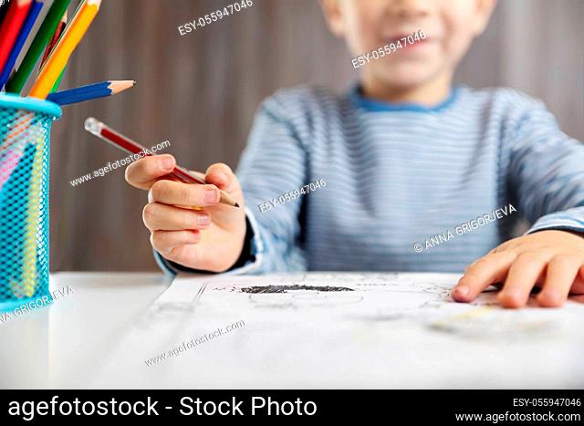 Little child at home drawing on the paper. Boy sitting on the chair at table. Five years old kid writing at desk in room with wooden background