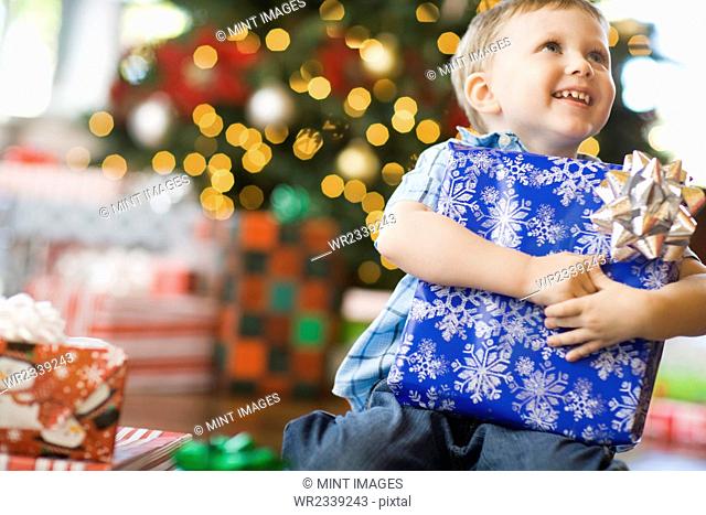 A young boy by a Christmas tree hugging a large wrapped present