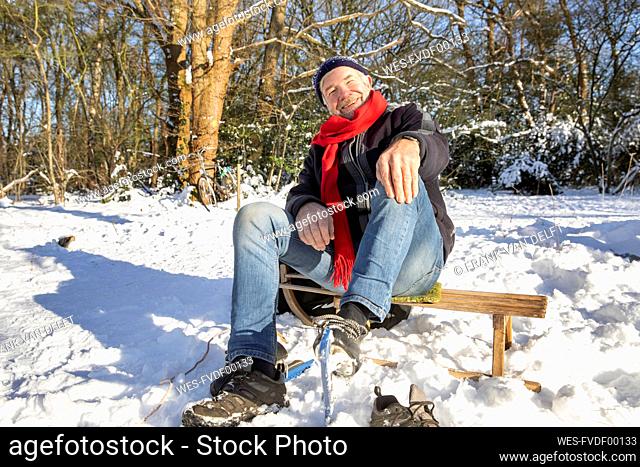 Happy senior man sitting at sled on snow during sunny day