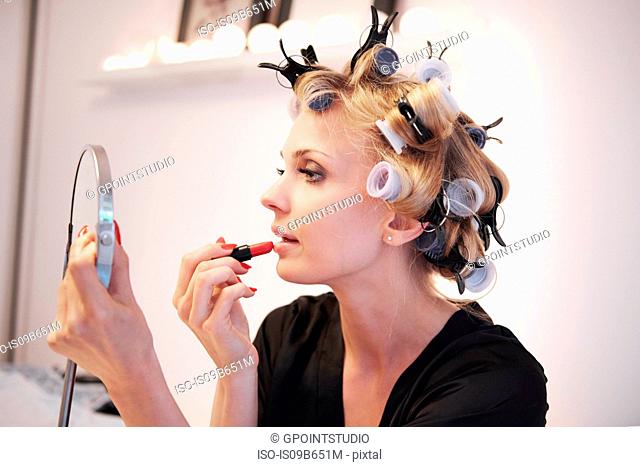 Woman with hair rollers applying lipstick in mirror
