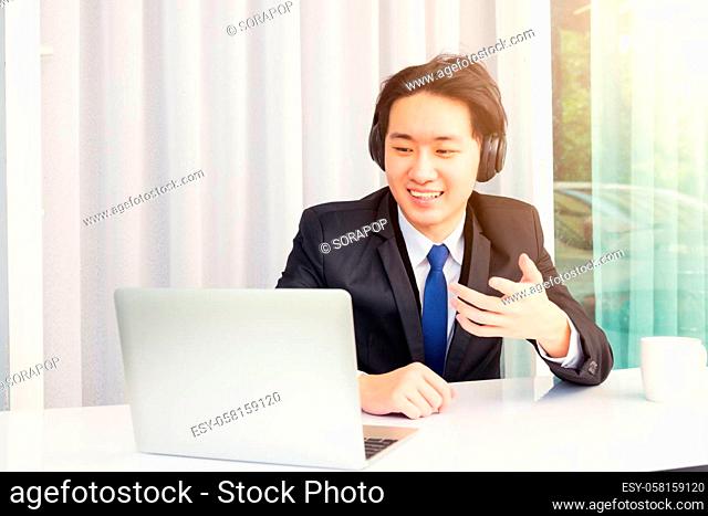 Work from home, Asian young businessman smile wearing headphones and suit video conference call or facetime on desk and raise his hand to explain to colleagues...