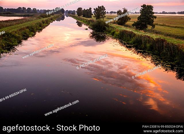 16 August 2023, Mecklenburg-Western Pomerania, Friedrichsmoor: The clouds colored orange-red by the rising sun are reflected in the water of the Elde River in...