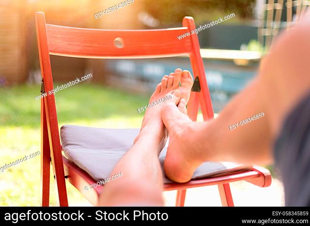 Young man is relaxing at home. Resting feet on a red chair, own garden in the blurry background