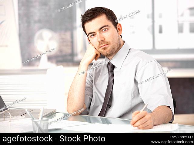 Young businessman working in bright office, sitting at desk, writing notes, thinking