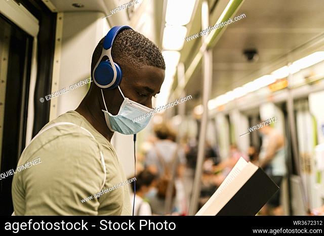 Close-up of young man wearing mask reading book and listening music through headphones in train