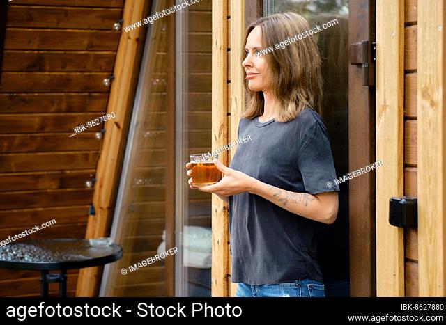 Cheerful young woman leaning against glass door of triangular wooden cabin with a cup of herbal tea
