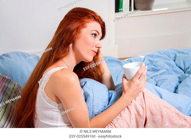 young red-haired woman sitting in the morning after getting up on her bed with a cup of coffee