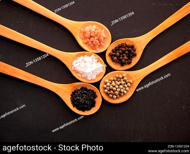 Wooden spoons with himalayan salt, black hawaii salt, common salt, salt flakes and peppercorns on a slate plate