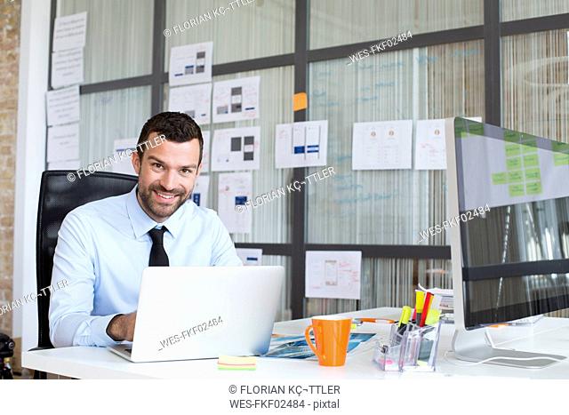 Portrait of smiling businessman in office using laptop at desk