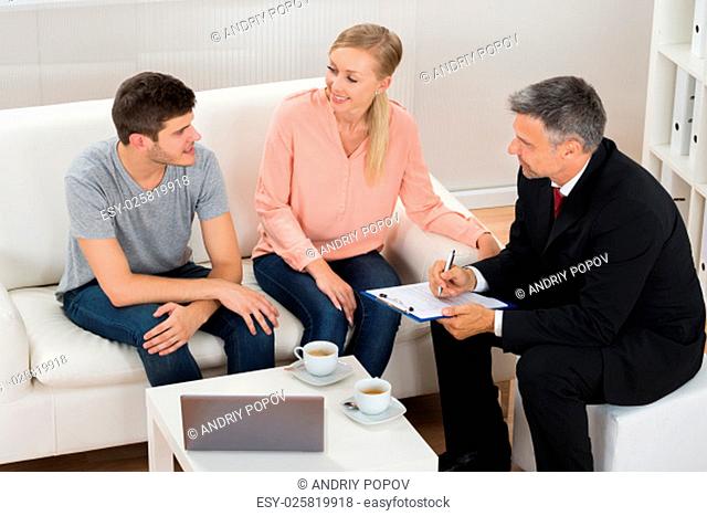 Real Estate Agent Talking To Young Couple Sitting On Sofa
