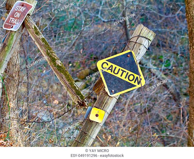 A fence falling into a ravine appears broken and tilted with a stay on trail sign and a bright yellow caution sign