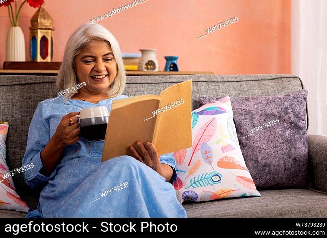 Cheerful old woman drinking tea and reading a book at home