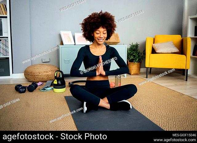 Smiling woman sitting cross-legged with eyes closed on exercise mat in living room