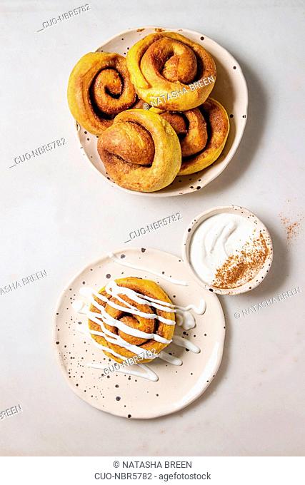 Homemade pumpkin cinnamon bun rolls sweet autumn baked dessert with cream cheese sauce in spotted ceramic plates over white marble background
