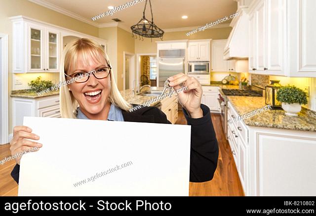 Happy young woman holding blank sign and keys inside beautiful custom kitchen