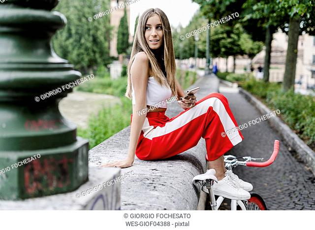 Smiling teenage girl with cell phone sitting on a wall at the riverside next to bicycle