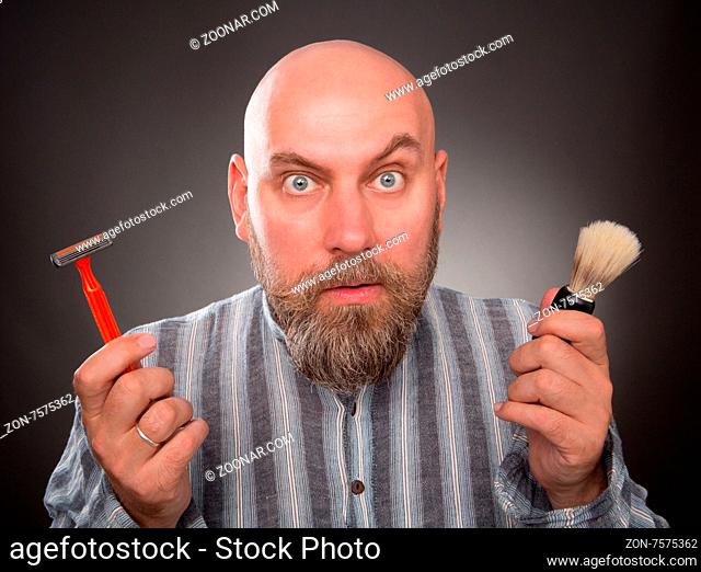Astonished jailbird posing with razor and brush in his hands. Bold bearded man is very surprised isolated on dark grey