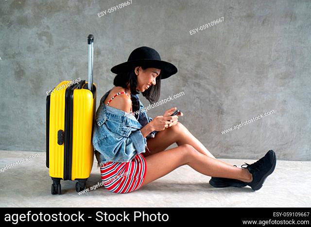 Portrait of smiling young Asian woman with luggage and mobile phone isolated over concrete wall. People lifestyle, travel concept