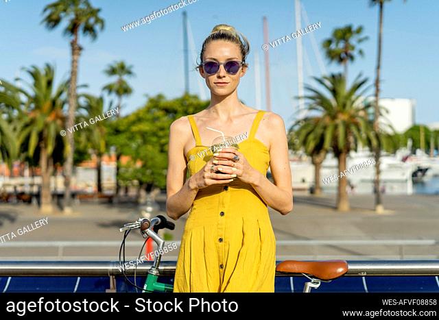 Beautiful woman with bicycle wearing sunglasses holding smoothie in city