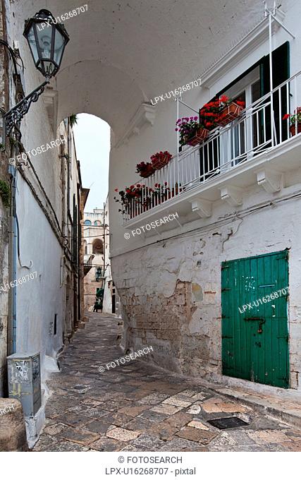 Street in Ostuni, with typical white stucco walls, stone paving, wrought iron lamp, and archway, with flowers decorating, spring afternoon, Salento, Puglia