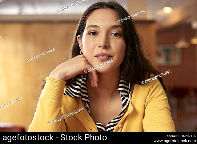 Portrait of young ethnic woman wearing yellow sweater with black and white striped blouse sitting at bar in kitchen of downtown loft