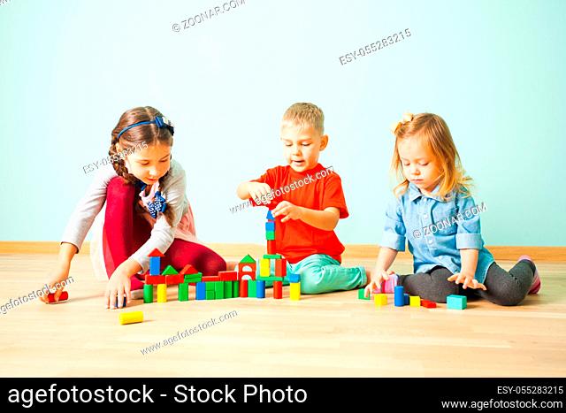 Three friends sitting on a wooden floor building from colorful blocks. Two pretty girls helping younger blond boy to create a tower from colorful bricks