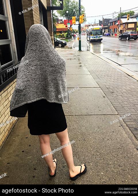 A young woman in black dress with a make-shift rain cover waits for a city bus in the rain, Ontario, Canada