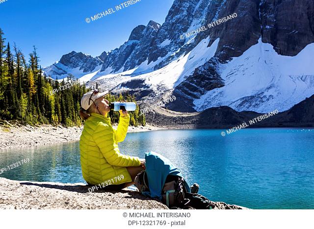 Female hiker drinking from water bottle on rocky alpine lake shoreline with snowed mountain cliffs in the background; Vermillion Crossing, British Columbia