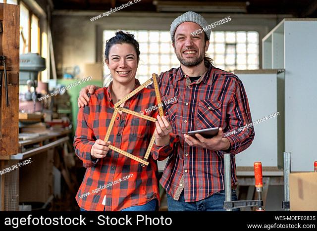 Portrait of happy craftswoman and craftsman holding tablet and yardstick in their workshop