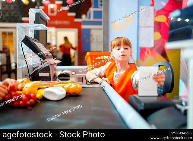 Cute girl in uniform at the cash register makes a check for the purchase, saleswoman, playroom. Kids plays sellers in imaginary supermarket