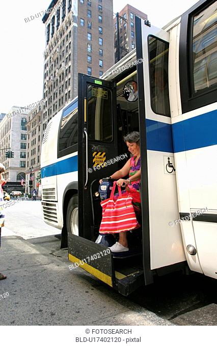 Woman with a disability (carrying a bright pink striped tote!) exiting a city bus via a lift