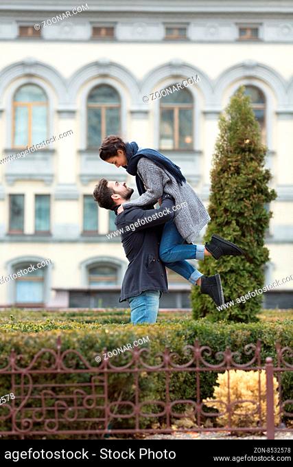 Happy and astonished couple enjoying the day in the autumn park. Handsome man lifting his girl-friend and smiling