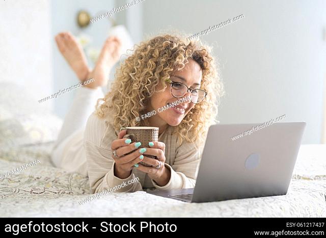 Smiling woman in eyeglasses and curly hair watching media content using laptop on comfortable bed at home. Happy woman watching movie on laptop while drinking...