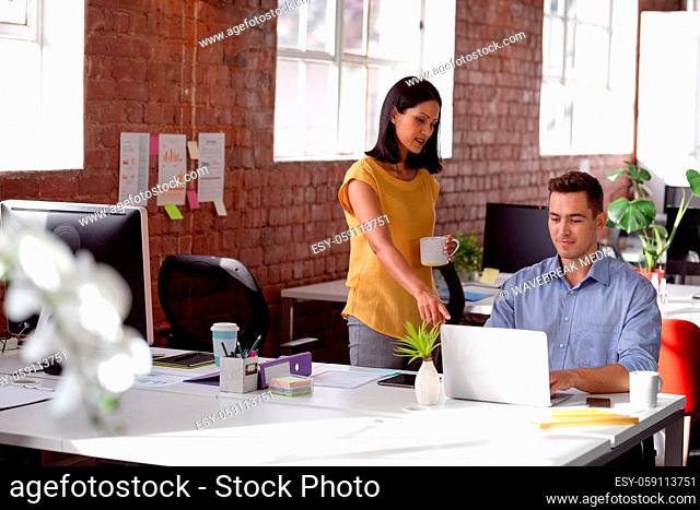 Caucasian male and female colleague in discussion at desk looking at laptop and drinking coffee