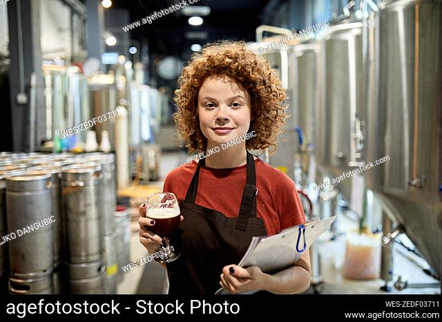 Portrait of confident woman holding clipboard and beer glass in craft brewery